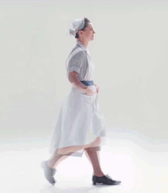 A person is walking to the right against a plain white background. They are wearing a light blue and white dress with short sleeves, a blue belt, and black shoes. They also have a white headscarf on. Their hands are in the pockets of the dress, resembling an NHS promotional video theme.