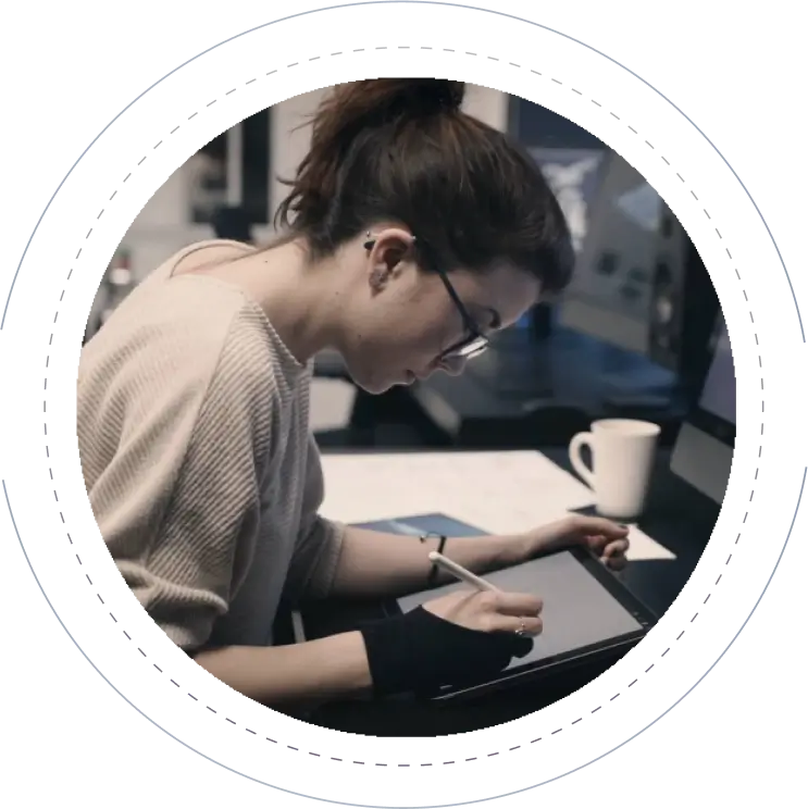 A person with glasses and tied-back hair is working on a tablet using a stylus. They are seated at a desk with a computer monitor and a white coffee mug in the background. They are wearing a light-colored sweater and a hand brace.