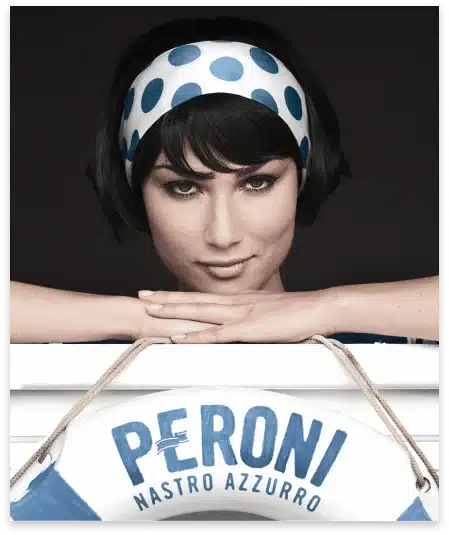 A Woman With Short Dark Hair And A Blue And White Polka Dot Headband Rests Her Chin On Her Hands. In Front Of Her Is A Life Preserver With The Text &Quot;Peroni Nastro Azzurro.&Quot; The Background Is Black, Highlighting Her Face And The Life Preserver. Meet The Dragonfly Team! A woman with short dark hair and a blue and white polka dot headband rests her chin on her hands. In front of her is a life preserver with the text "PERONI NASTRO AZZURRO." The background is black, highlighting her face and the life preserver. Meet the Dragonfly Team!