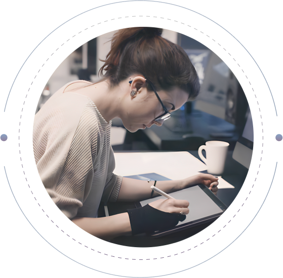 A person with glasses and dark hair tied in a ponytail works on a digital drawing tablet. They are seated at a desk with a computer monitor and a white coffee mug nearby. The scene appears to be in a modern, well-lit workspace.