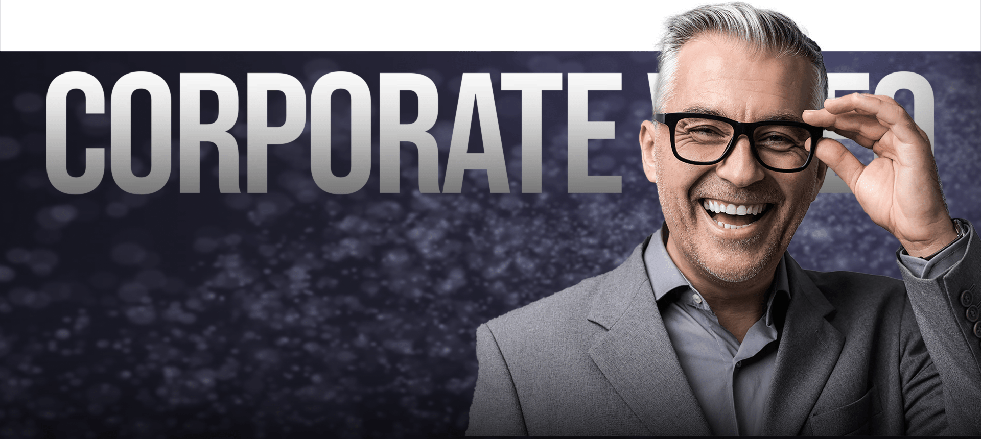 A Smiling Man With Gray Hair And Glasses, Wearing A Gray Suit, Stands In Front Of A Backdrop With The Large Text &Quot;Corporate Video&Quot; Partially Visible. The Background Features A Dark, Blurred Bokeh Effect. A smiling man with gray hair and glasses, wearing a gray suit, stands in front of a backdrop with the large text "CORPORATE VIDEO" partially visible. The background features a dark, blurred bokeh effect.