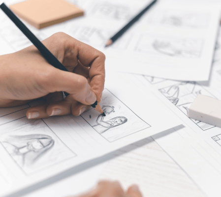 A Person'S Hands Are Drawing A Storyboard On White Paper Using A Pencil, Contemplating Animation Vs Live-Action. The Desk Is Cluttered With Sketches, An Eraser, And Sticky Notes. The Storyboard Includes A Rough Sketch Of A Character'S Face In One Of The Frames, Considering Different Video Styles. A person's hands are drawing a storyboard on white paper using a pencil, contemplating Animation Vs Live-Action. The desk is cluttered with sketches, an eraser, and sticky notes. The storyboard includes a rough sketch of a character's face in one of the frames, considering different video styles.