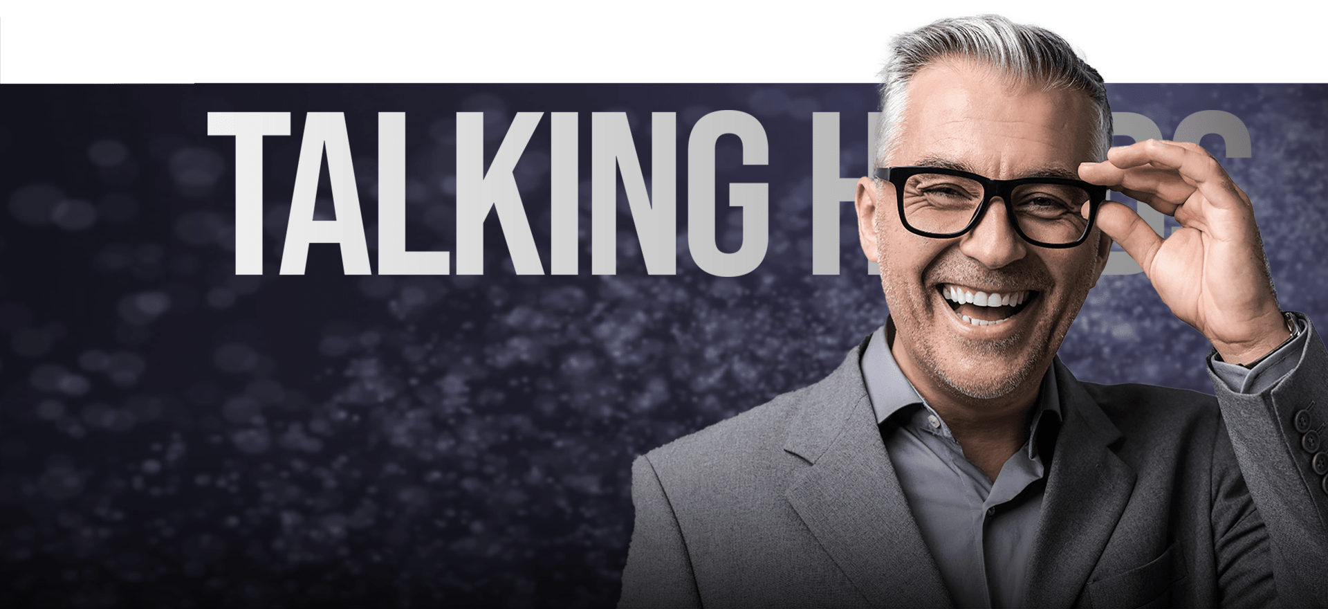 A Middle-Aged Man With Gray Hair And Glasses Is Smiling Broadly While Adjusting His Glasses. He Is Wearing A Gray Suit And Stands In Front Of A Dark, Blurred Background With White Specks And The Words &Quot;Talking Heads&Quot; In Large, Bold Letters. A middle-aged man with gray hair and glasses is smiling broadly while adjusting his glasses. He is wearing a gray suit and stands in front of a dark, blurred background with white specks and the words "TALKING HEADS" in large, bold letters.