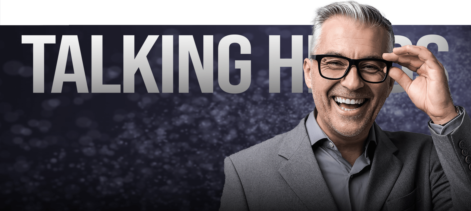 A smiling man with gray hair and glasses is adjusting his glasses while wearing a gray suit and shirt. Behind him is a backdrop with a subtle pattern and large text reading "TALKING HEADS." The overall mood is professional and approachable.