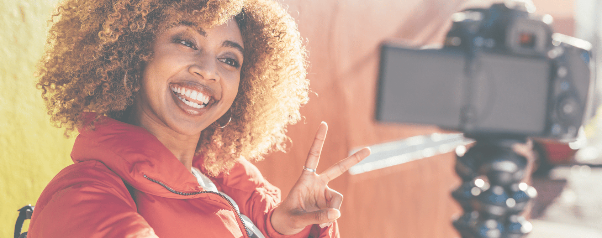 A person with curly hair wearing a red jacket smiles brightly and makes a peace sign while looking at a camera on a tripod. The background is a gradient of yellow and orange hues, creating a vibrant atmosphere perfect for a social media video.