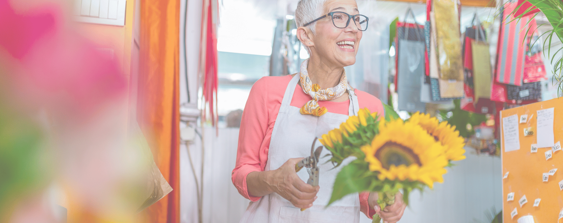 A smiling elderly person with short gray hair and glasses, wearing an apron over a coral top and a scarf, holds a bouquet of sunflowers in a vibrant and colorful flower shop. The background, bustling with plants and hanging decorations, showcases the charm that draws in small businesses for unique video production opportunities.